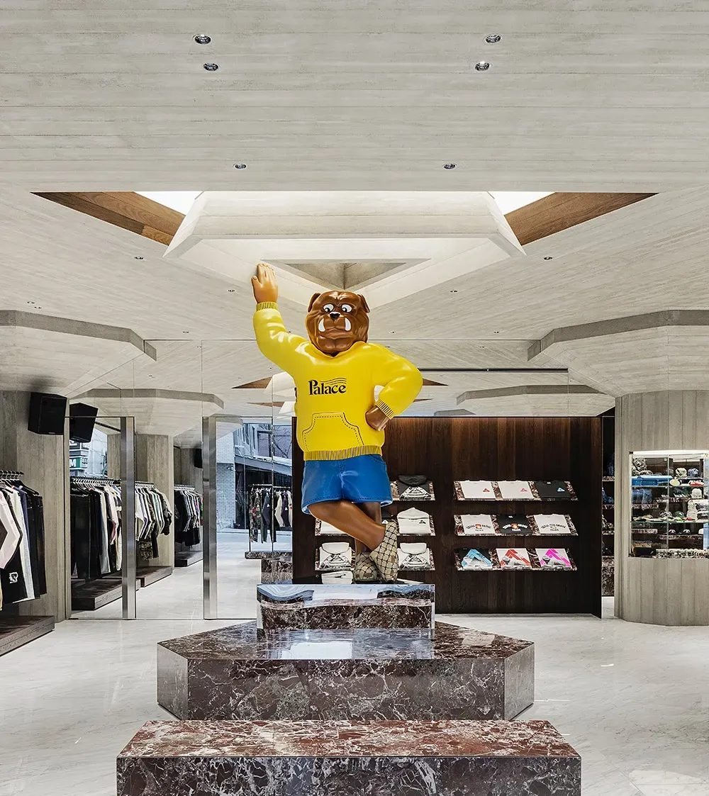 Front view of a giant bulldog figure in a yellow Palace hoodie on a marble plinth, with T-shirt shelves and clothing rails