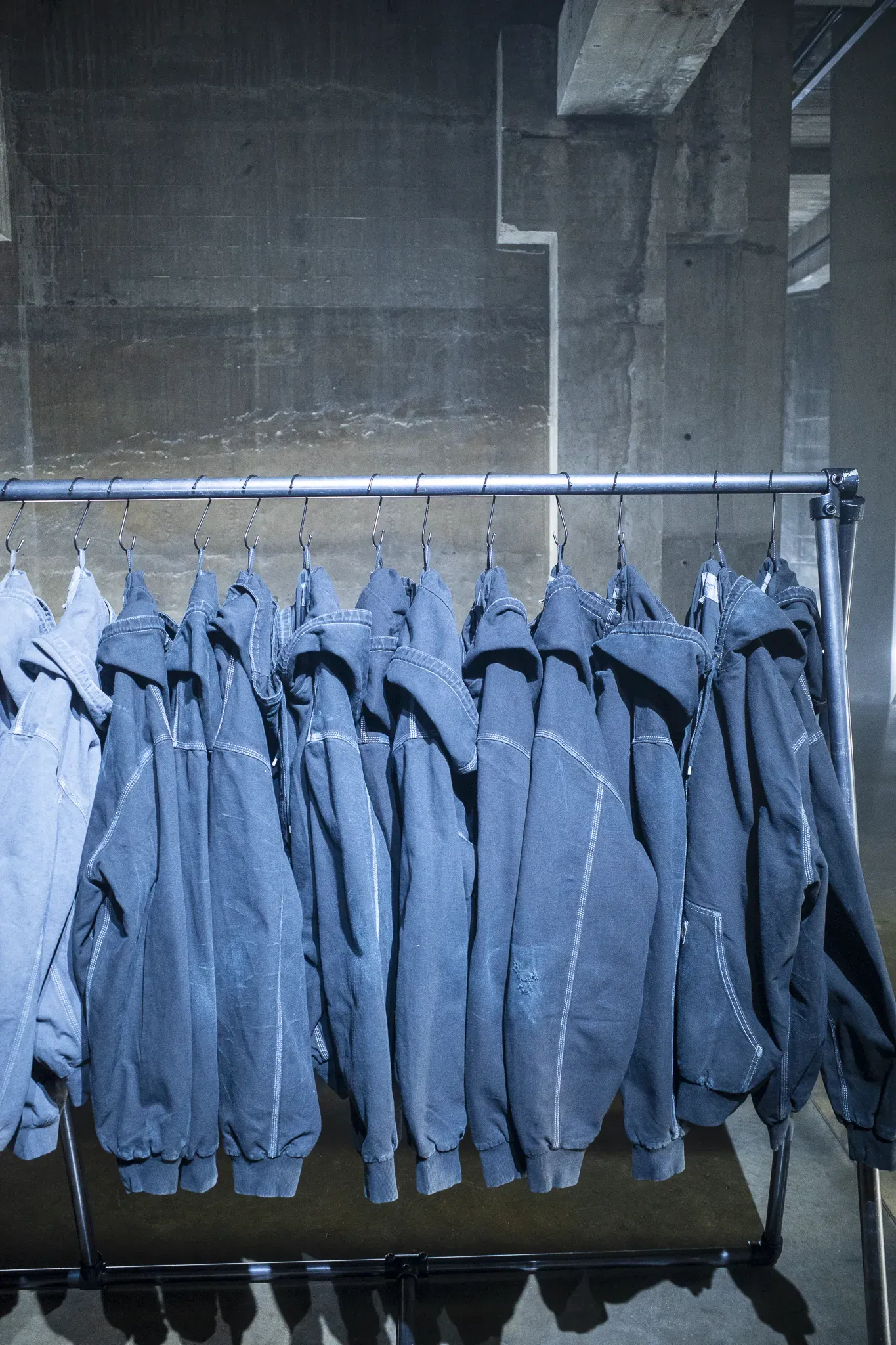 Row of faded blue hooded work jackets hanging on S-hooks from a metal rail against a raw concrete wall
