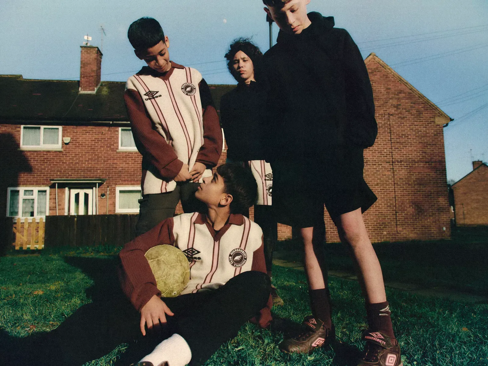 Four kids in Umbro and Highsnobiety knitwear and boots pose on grass before brick houses, one sitting with a worn ball