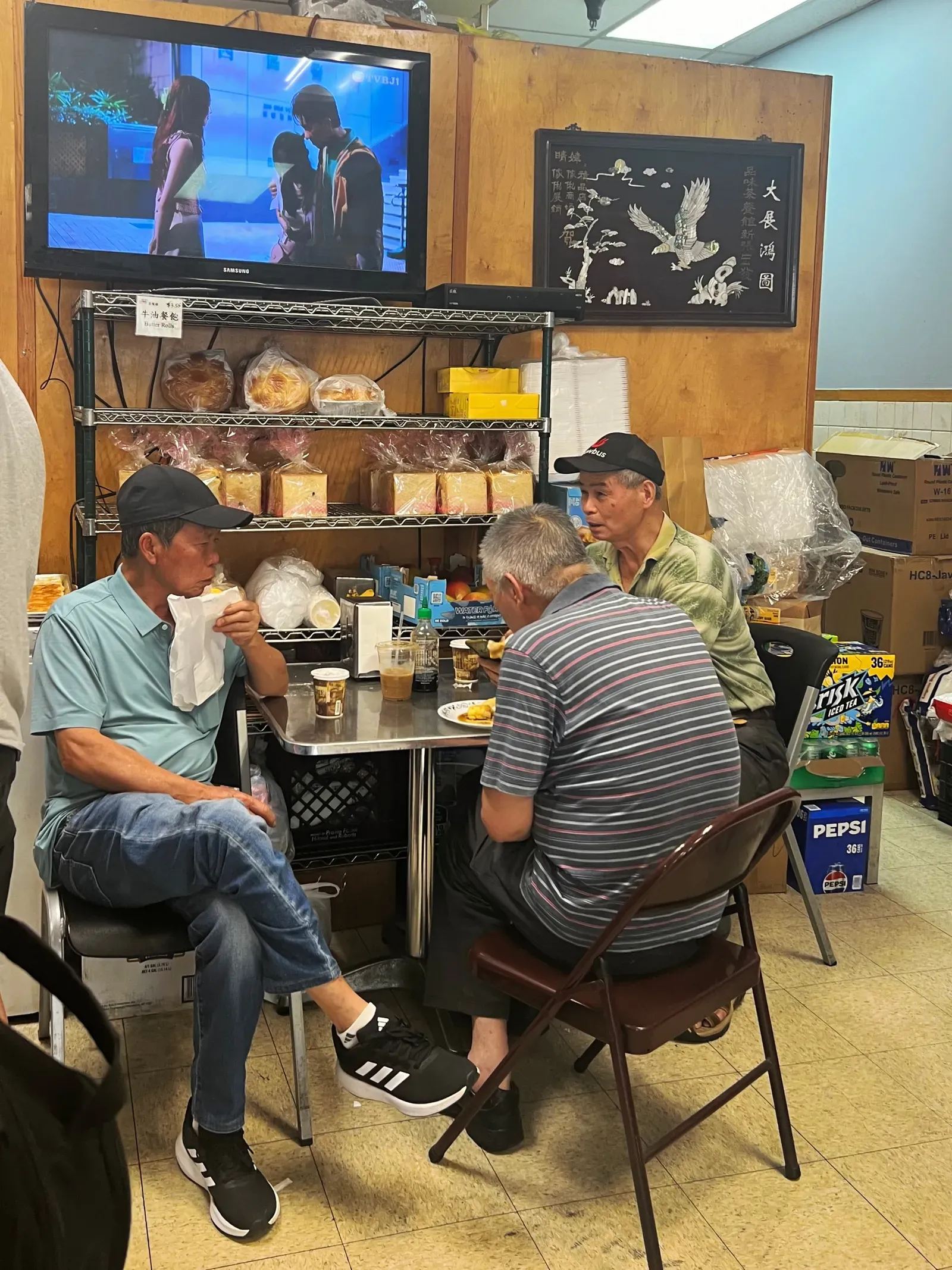 Three older men eating and talking at a small steel table in a Chinese bakery café beneath a TV