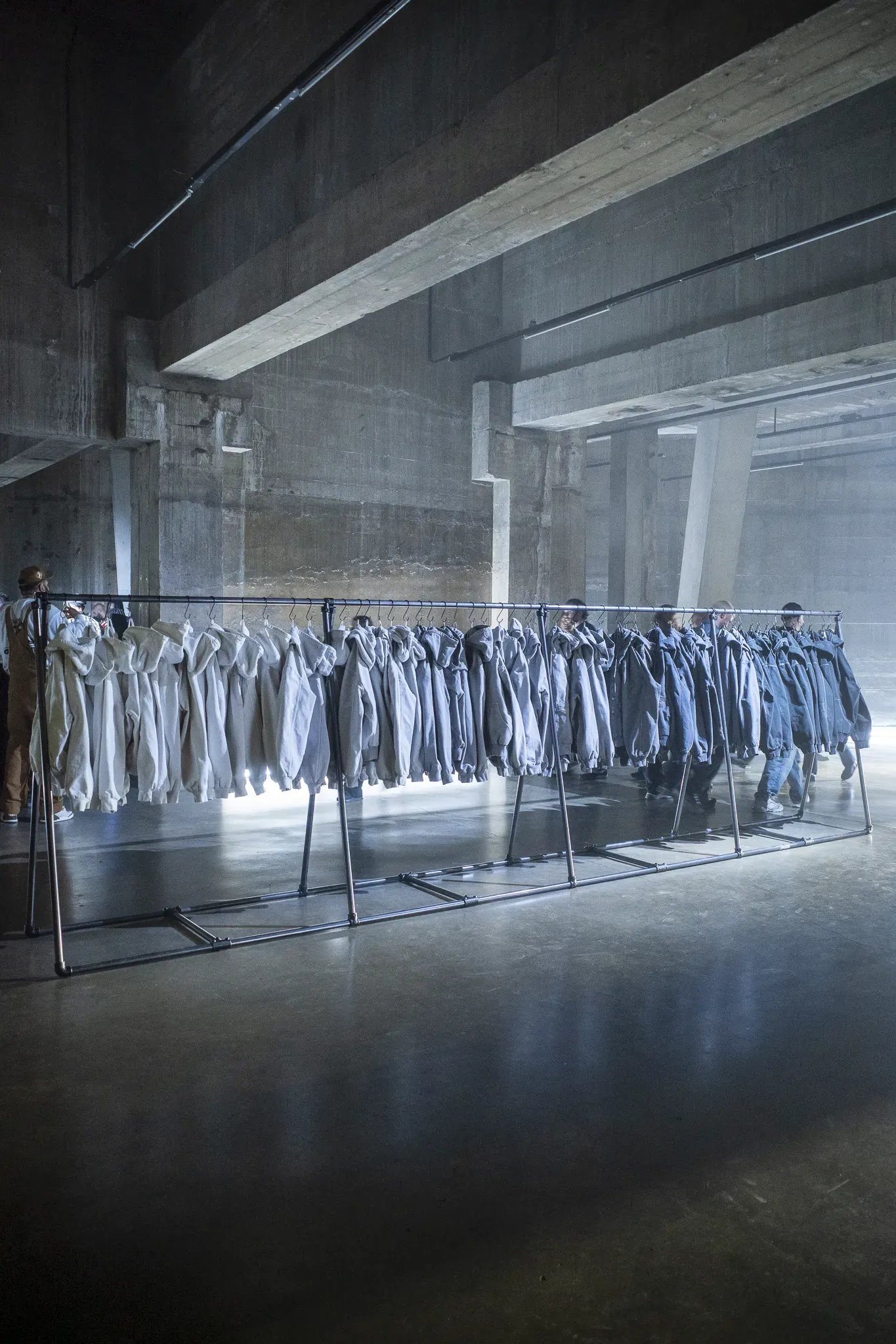 Long pipe rail of hooded jackets shading from off-white to faded blue in a concrete hall, guests walking behind