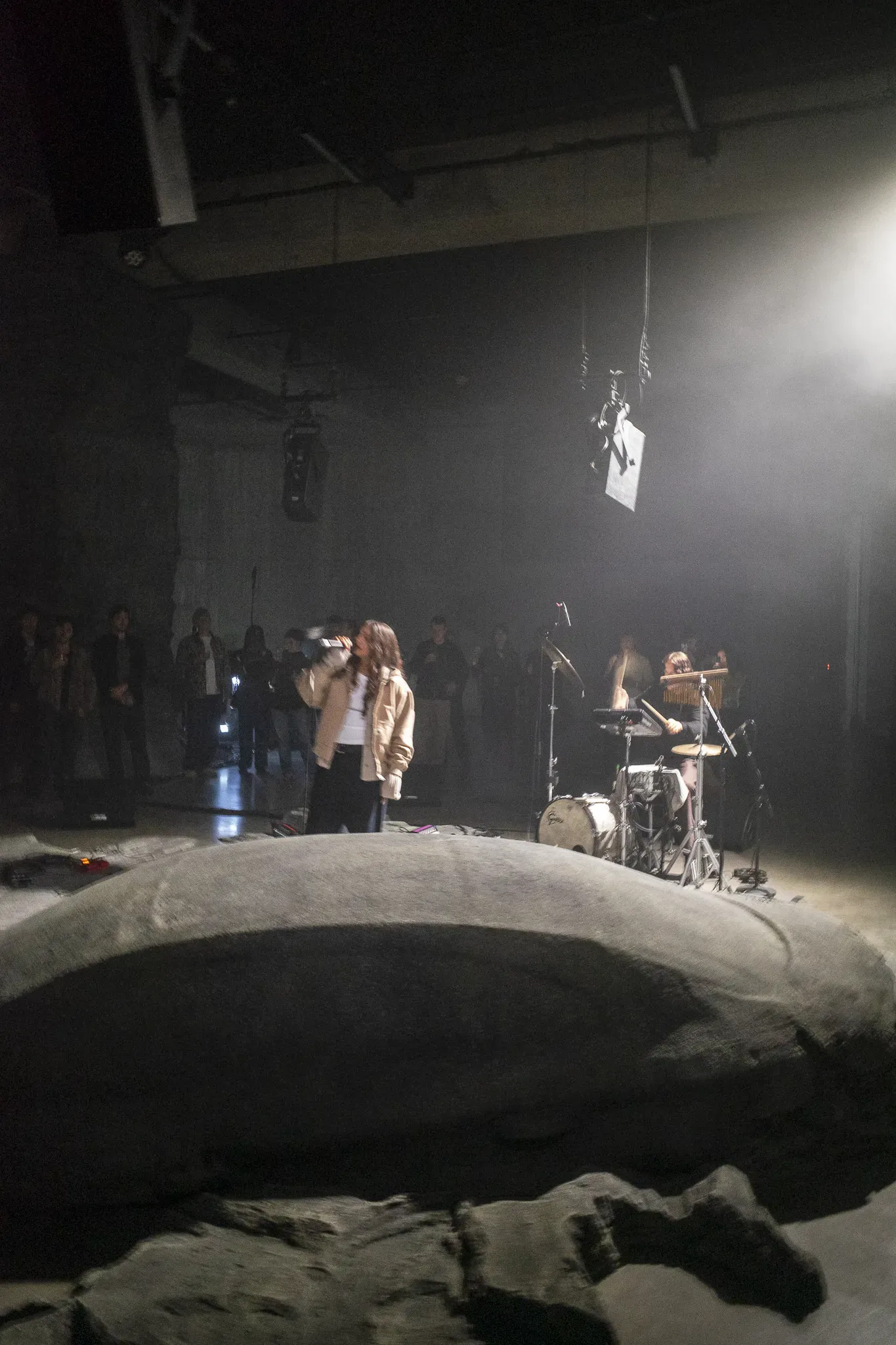 Singer in a beige jacket performs with a drummer beside a rock-like sculpture as guests watch in the dark