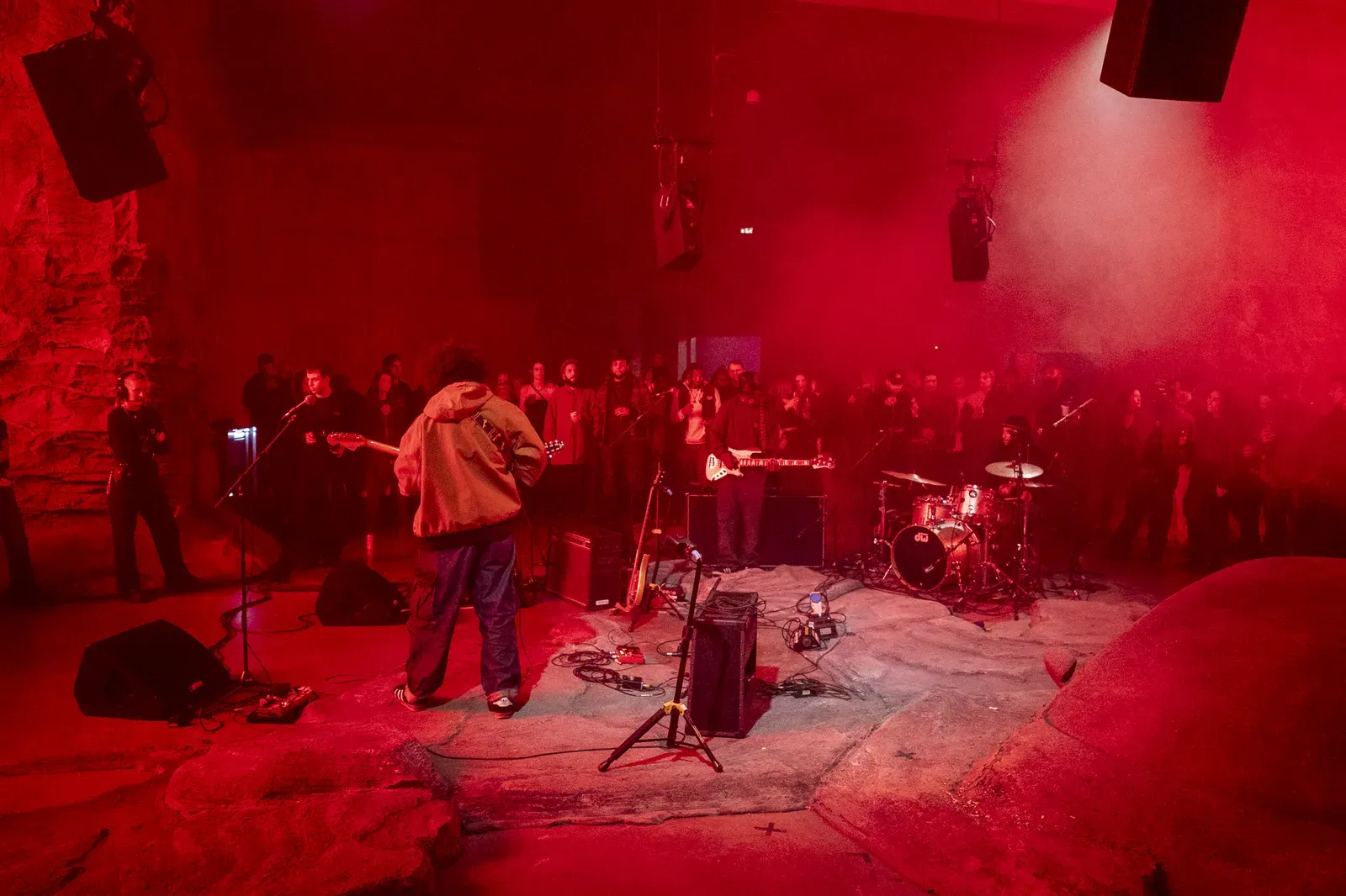 Band plays on a rock-like stage bathed in red light, a guitarist in a hooded jacket facing the packed crowd