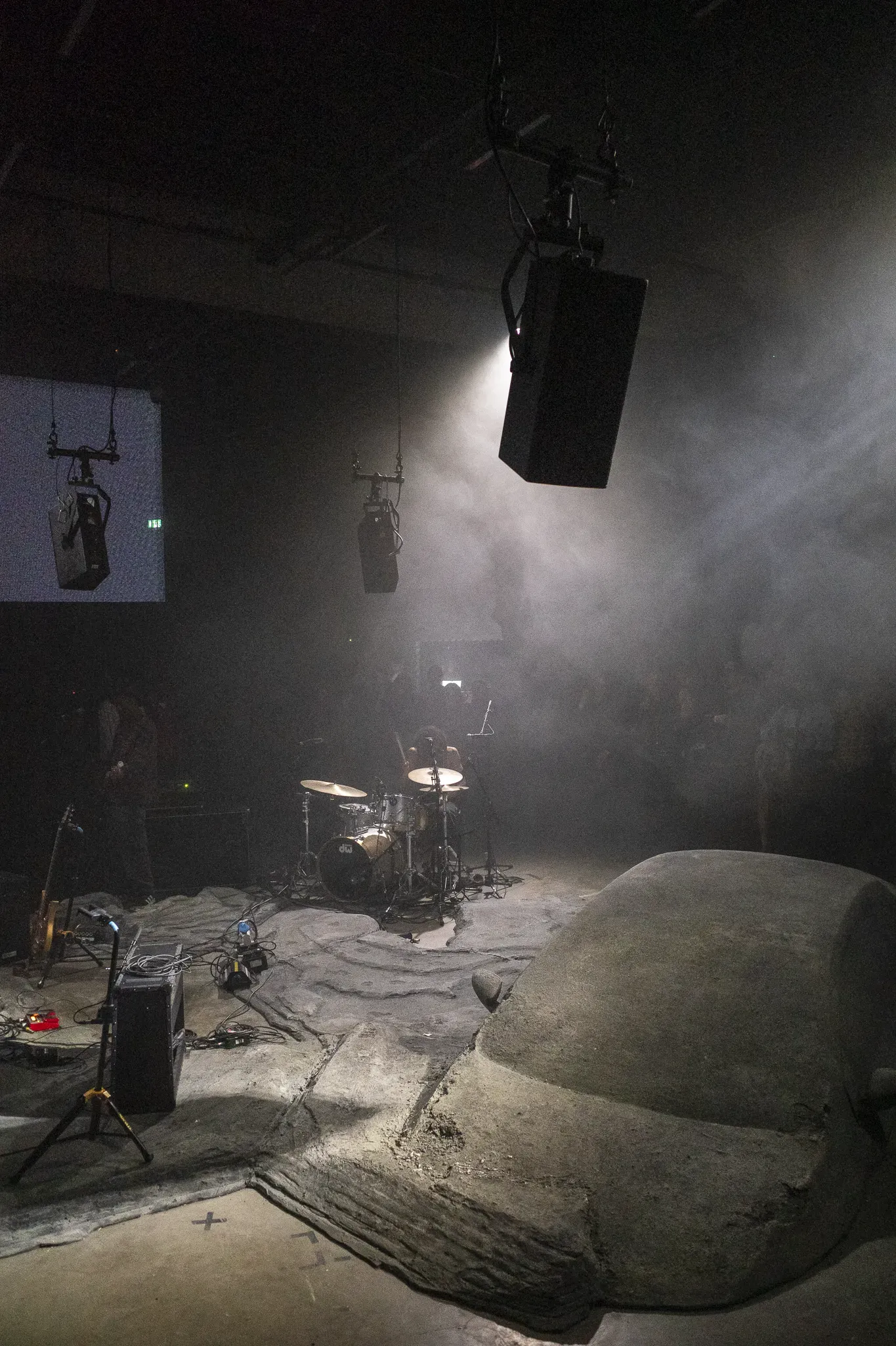 Drummer plays in thick haze under hanging speakers, a large rock-like sculpture in the foreground