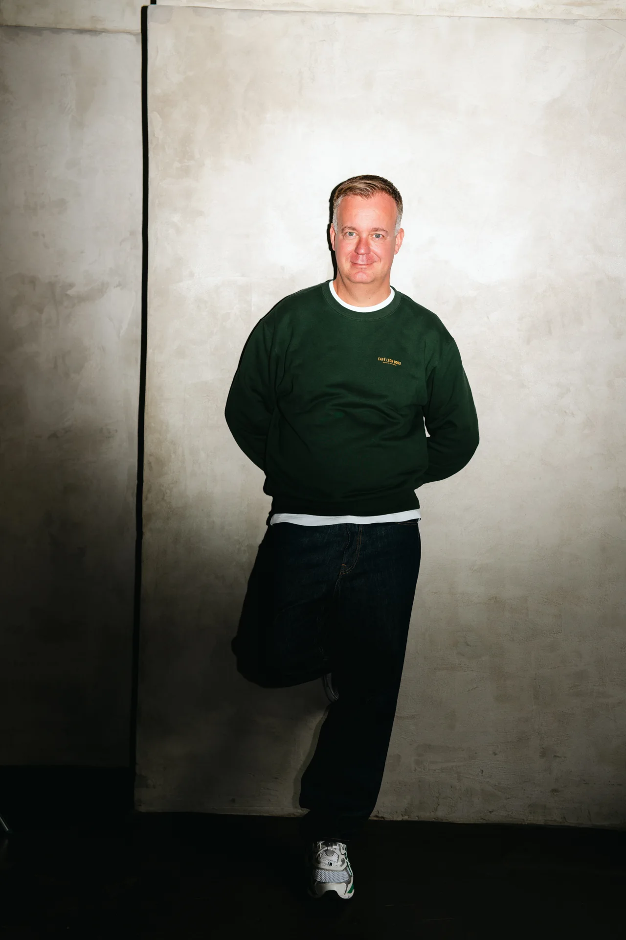 Jürgen Alker standing in a green sweatshirt against a pale architectural wall
