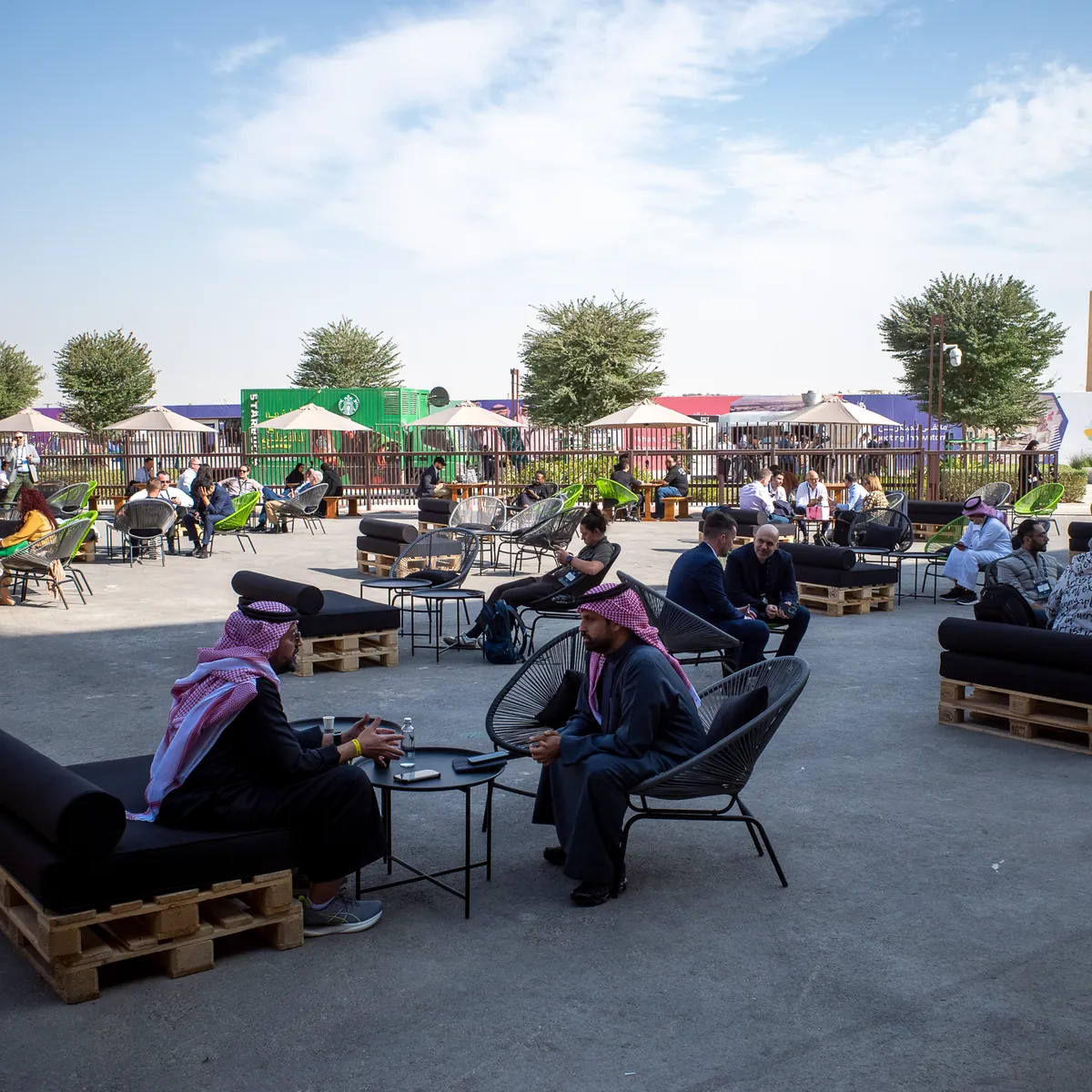 Men in thobes sitting in low chairs in an open-air seating area.